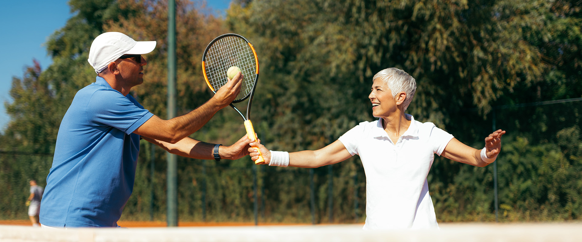 Ein Tennistrainer in blauem Shirt und weißer Kappe assistiert einer Spielerin in weißem Poloshirt bei der Handhabung von Tennisball und Schläger auf einem Sandplatz des Tennisclubs Schwarz-Weiß Montabaur.