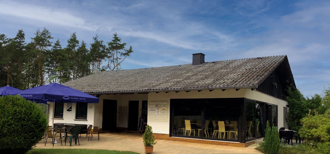 Das Clubhaus des Tennisclubs Schwarz-Weiß Montabaur mit Sonnenterrasse, Outdoor-Sitzgelegenheiten und großem Glasfrontbereich, umgeben von Bäumen und Grünflächen.