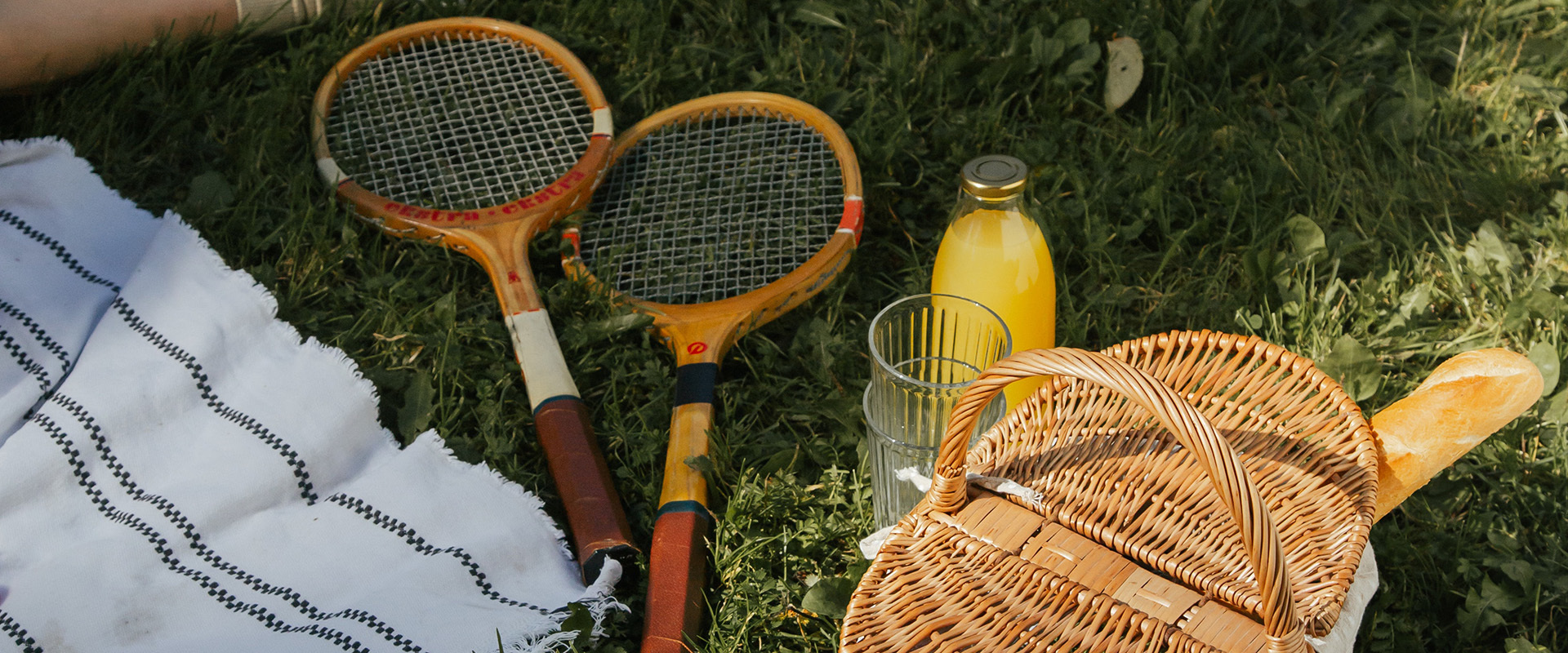 Zwei alte Tennisschläger, ein Picknickkorb mit Orangensaft, einem Glas und einem Baguette sowie eine gestreifte Decke liegen auf einem Grasrasen – ein Bild, das die traditionelle und gemütliche Atmosphäre des Tennisclubs Schwarz-Weiß Montabaur e.V. einfängt.
